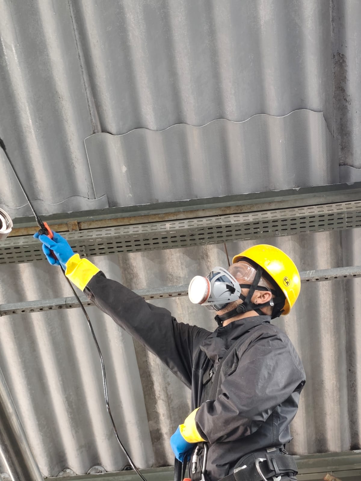 trabalhador com capacete amarelo, máscara respiratória e luvas azuis pulverizando tratamento em telhado de tel
